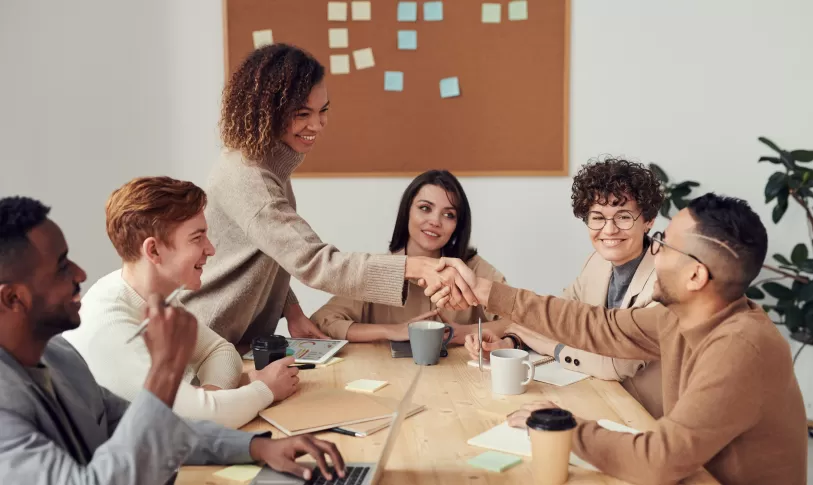 Een groep mensen die rond de tafel zit en twee mensen schudden elkaar de hand