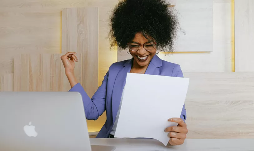 Vrouw achter laptop met papier in haar hand, ze kijkt heel erg blij