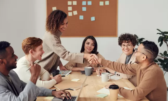 Een groep mensen die rond de tafel zit en twee mensen schudden elkaar de hand